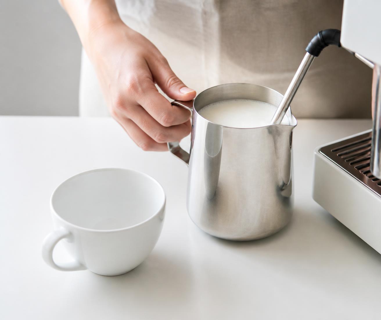 Barista steaming milk in a stainless jug next to a white cup